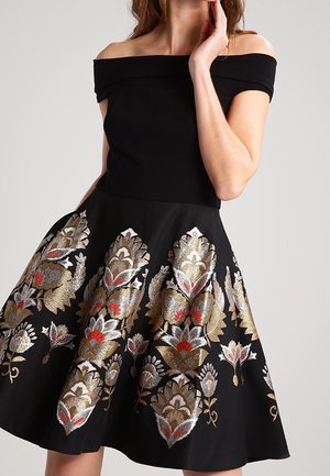 Woman wears off-shoulder black dress with flared skirt featuring large gold, silver, and red floral embroidery patterns.