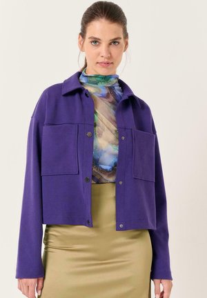 Lila, kurze Jacke aus weichem Stoff mit Kragen, zwei großen Fronttaschen und schwarzen Druckknöpfen.
