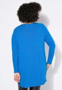 Langärmliges Oberteil in strahlendem Blau, mit rundem Ausschnitt, abgerundetem Saum und einer leichten, glatten Textur. Darunter werden schwarze Leggings getragen.