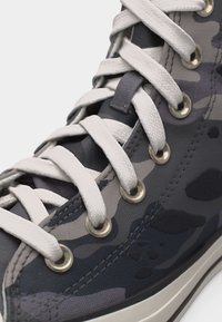 Ψηλό παπούτσι αθλητικό με γκρι και μαύρο καμουφλάζ σχέδιο, λευκά κορδόνια και μεταλλικά ρύγχη, σε κοντινή λήψη της μπροστινής πλευράς.