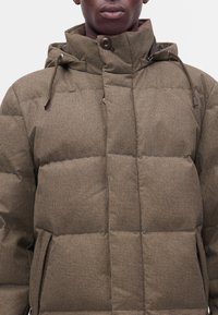 Gros plan d'une personne portant une doudoune marron à carreaux avec un col montant fermé par un bouton et des cordons sur la capuche.