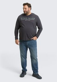 Grijze longsleeve shirt met witte en groene tekst, gecombineerd met blauwe jeans en zwarte schoenen. Model staat met één hand in de zak.