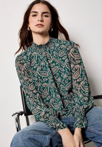 Grüne, gemusterte Bluse mit gerafftem Halsausschnitt und langen Ärmeln, die florale Motive in Creme und Pink auf einem dunklen Hintergrund zeigt.