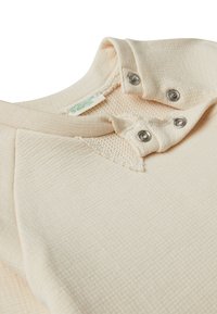 Top a maniche lunghe color beige chiaro in tessuto testurizzato, con scollatura rotonda, bottoni a pressione e un'etichetta con la scritta "United Colors of Benetton."