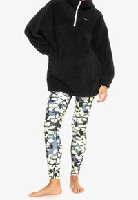 Zwarte fleece trui met capuchon en rits aan de voorkant, gecombineerd met meerkleurige bloemenleggings met blauw- en crèmeachtige patronen.