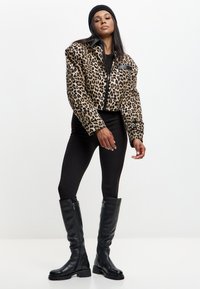Donna che indossa una giacca maculata, leggings neri, berretto nero e stivali neri fino al ginocchio, in posa su sfondo bianco.