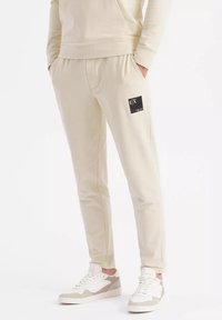 Beige Sweatpants mit einem elastischen Bund, Seitentaschen und einem schwarzen Etikett am Oberschenkel. Kombiniert mit weißen und beigen Sneakers.