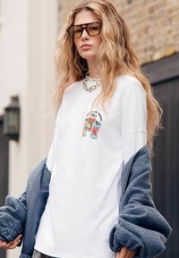 Fehér, túlméretezett póló színes üdítős doboz grafikával, hozzá társítva kék, bolyhos dzseki, ezüst lánc nyaklánc és nagy napszemüveg.