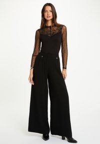 Top negro con encaje y mangas transparentes con puntos, combinado con pantalones negros de pierna ancha. Textura suave con mínimos acentos plateados en la cinturilla.