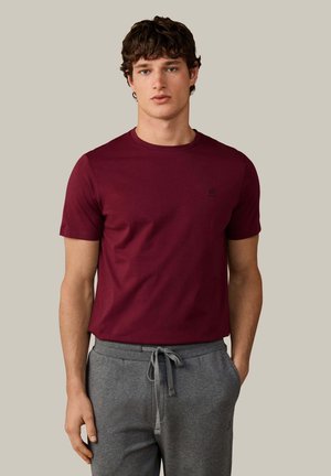 Jonge man met kort krullend haar, gekleed in een effen bordeauxrood T-shirt en grijze joggingbroek met trekkoord, staand tegen een neutrale achtergrond.