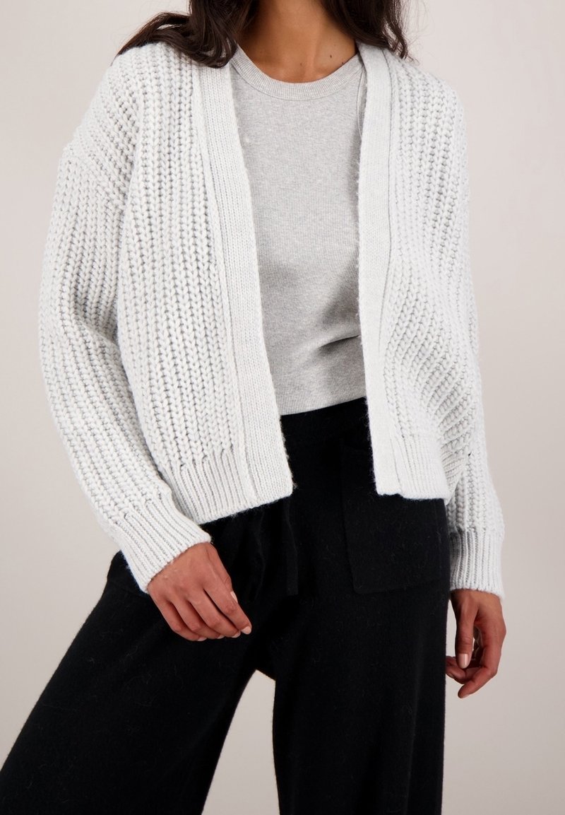 Cárdigan de punto gris claro con frente abierto, puños acanalados y patrones texturizados; llevado sobre una camisa gris a juego y pantalones negros.