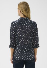 Femme aux longs cheveux blonds vue de dos, portant une chemise à boutons florale bleu marine à manches retroussées et un jean foncé.