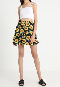 Vrouw in een witte cropped tanktop, een knoopsluiting marineblauwe rok met gele zonnebloemprint en zwarte slip-on sandalen, staande tegen een witte achtergrond.