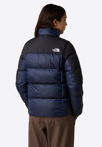 Kvinna med mörkt hår som bär en marinblå och svart pufferjacka med The North Face-logotyp, stående vänd bort från kameran mot en enfärgad bakgrund.