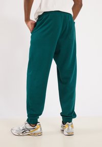 Teal sweatpants with a relaxed fit, elastic waistband, side pockets, and ribbed cuffs. Paired with silver and gold athletic shoes.