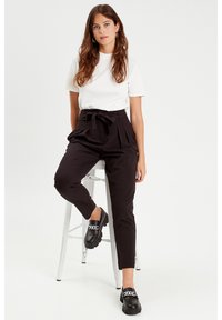 Weißes, kurzärmliges Oberteil, hochgeschnittene schwarze Hose mit Gürtelbindung und schwarze Slipper mit silberner Kettendetails. Model sitzt auf einem Hocker.