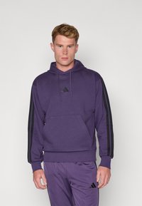 Sudoeste roxo com riscas pretas nas laterais, bolso canguru, capuz com cordão e um pequeno logotipo preto no peito. Tecido texturizado, corte largo.