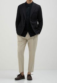 Navy blazer over a dark button-up shirt, paired with light beige tapered trousers and brown lace-up shoes, on a neutral background.