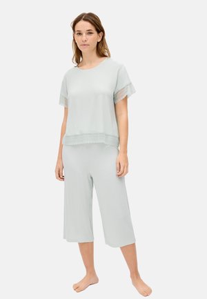 Mujer con blusa de manga corta gris claro con borde de encaje y pantalones anchos recortados a juego, de pie descalza sobre un fondo liso.