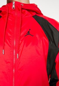 Veste en nylon rouge avec capuche, panneaux latéraux noirs et logo Jumpman noir sur la poitrine ; texture lisse avec fermeture éclair.