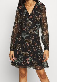 Schwarzes Blumenkleid mit V-Ausschnitt, langen transparenten Ärmeln, verziert mit einem Muster aus bunten Pflanzen und Blumen, strukturiertem Stoff, in der Länge oberhalb des Knies.