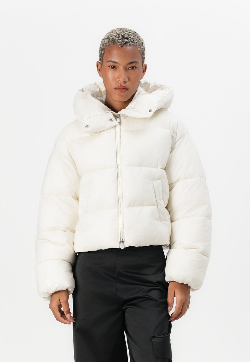 Korte witte pufferjas met een hoge kraag, grote capuchon en voorrits. Voorzien van een gewatteerd patroon en volumineuze mouwen.