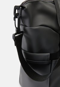 Saco preto, acolchoado, feito de material liso, com uma alça de ombro trançada e um bolso lateral, apresentando um fecho de correr e acessórios de plástico.