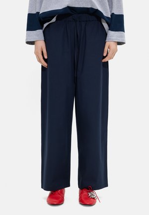 Osoba nosząca granatowe szerokie spodnie ze ściągaczem i jaskrawo czerwone buty z przyczepionymi srebrnymi dzwoneczkami, stojąca na białym tle.