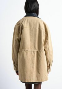 Femme portant un manteau beige oversize avec un col noir, debout de dos devant un fond blanc uni.
