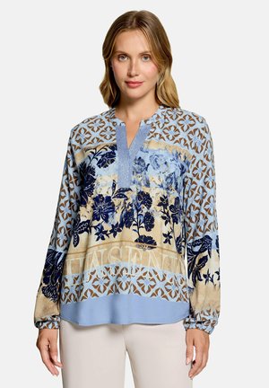 Frau mit schulterlangem blondem Haar, die eine langärmelige Bluse mit blauen und beigen Blumen- und geometrischen Mustern sowie helle Hosen trägt.