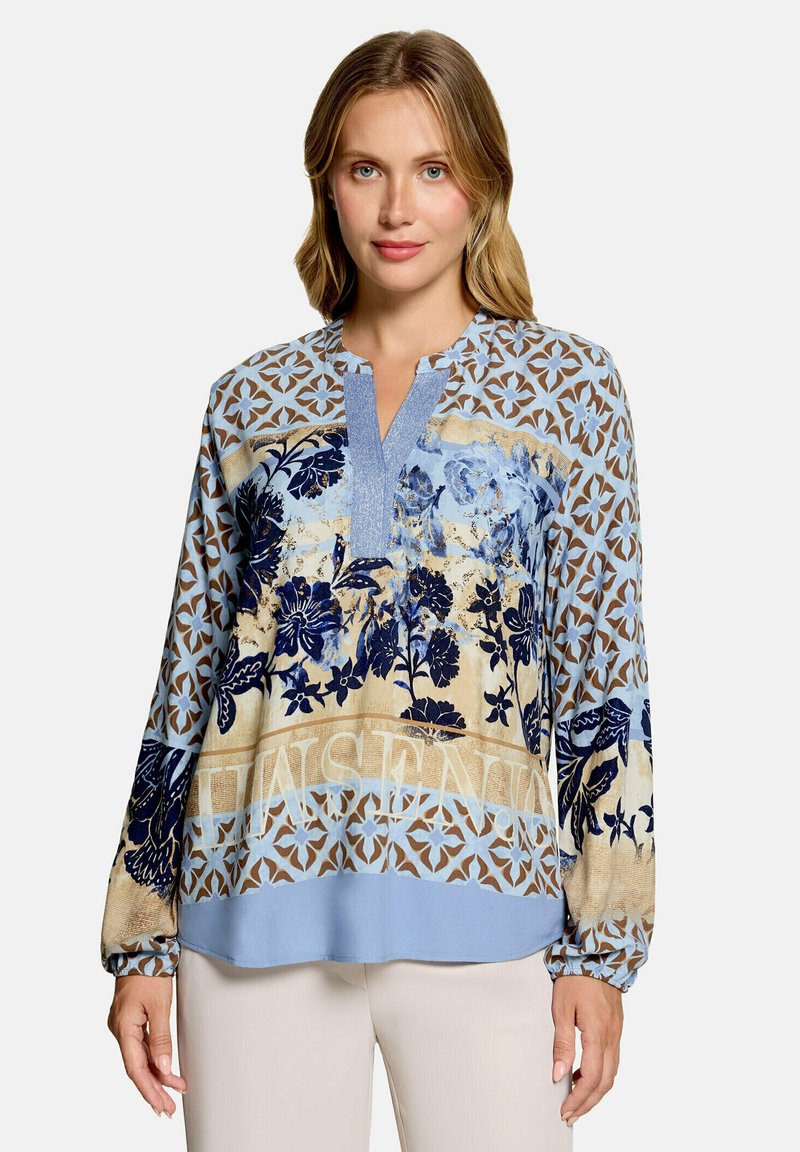 Femme aux cheveux blonds mi-longs portant un chemisier à manches longues avec des motifs floraux et géométriques bleus et beiges, et un pantalon clair.