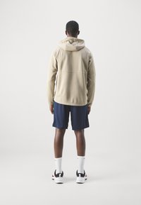 Beige hoodie med texturerad tyg, med en dragsko till huva, tillsammans med marinblå shorts. Modell med vita strumpor och svarta sneakers.