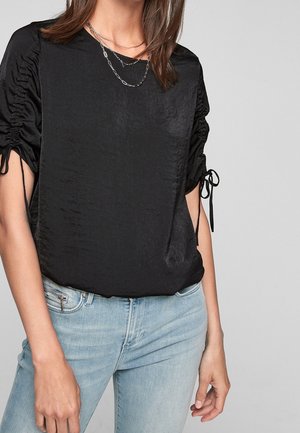Vrouw draagt een zwarte satijnen blouse met geplooide mouwen die met linten zijn vastgebonden en lichtblauwe spijkerbroek, tegen een effen lichte achtergrond.