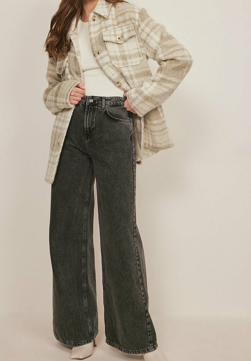 Geruite wollen jas in beige en wit, gecombineerd met zwarte high-waisted jeans met wijde pijpen. Model draagt een geribd wit topje en beige laarzen met hak.