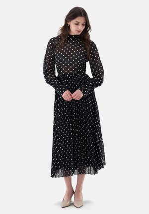Frau trägt ein schwarzes langärmeliges Midi-Kleid mit weißen Polka-Dots und beigen High Heels, steht mit verschränkten Händen und schaut nach unten.