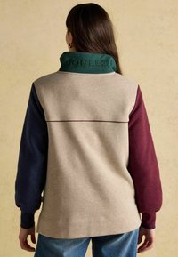 Žena v béžové fleecové bundě s námořnickým levým rukávem, burgundským pravým rukávem, zeleným límcem vyšitým nápisem "JOULES" a modrými džíny.