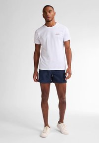 Bílé bavlněné tričko s malým červeným textovým logem, doplněné o tmavě modré plavky a béžové nízké tenisky. Neutrální pozadí.