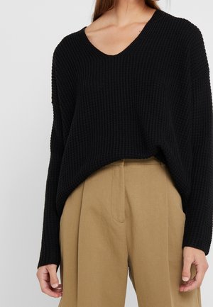 Person iført en sort langærmet striktrøje stoppet ned i beige vidde bukser med høj talje, vist fra nakke til hofter.