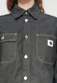 Γκρι τζιν πουκάμισο με μυτερό γιακά, μπροστινές τσέπες, κουμπιά με πατιλέτα και αντίθετες λευκές ραφές. Μπαλώμα με το λογότυπο της μάρκας στην αριστερή τσέπη.