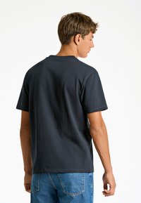 Tummansininen puuvillainen t-paita, jossa on lyhyet hihat, pyöreä pääntie ja rento istuvuus, yhdistettynä sinisiin farkkuihin. Minimalistinen muotoilu, ei näkyviä logoja.