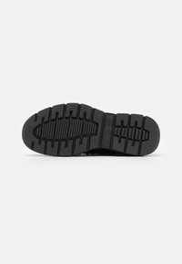 Black rubber shoe sole with a textured tread pattern, featuring deep grooves and ridges for traction. Flat, oval-shaped design.