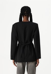Blazer noir ajusté à la taille cintrée et aux longues manches. Présente un col rond, une texture lisse et des accents de boutons sur les manches.