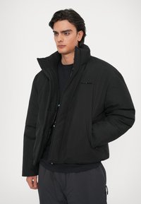 Uomo con un piumino nero con collo alto e mani in tasca, in piedi contro uno sfondo chiaro e uniforme.