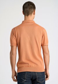 Kurzärmliges Poloshirt in hellorange, mit strukturiertem Strickmuster, Kragen und geripptem Saum. Getragen mit dunklen Jeans.