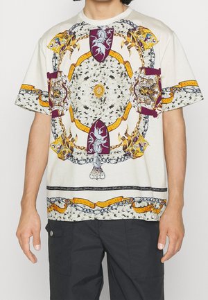 Kremowa koszulka z krótkim rękawem z misternymi wielokolorowymi wzorami, tarczami i łańcuchami. Odważne wzory i faktury na całym ubraniu.