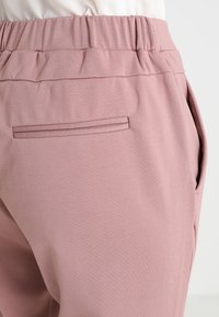 Pantalon en tissu doux rose avec un waistband froncé, des poches latérales et un élégant détail de poche cousue à l'arrière. Texture lisse.