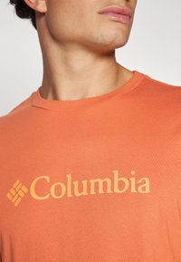 Narančasta pamučna majica s okruglim izrezom i istaknutim žutim logom "Columbia" na prsima, s glatkom i mekom teksturom.