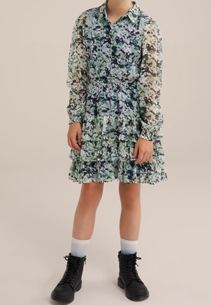 Barn iført en langærmet, blomsterprint kjole med flæser, hvide sokker og sorte snørestøvler, stående mod en ensfarvet baggrund.