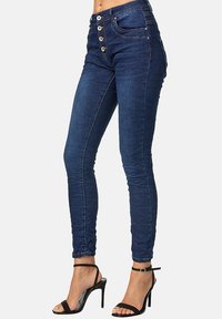 Jeans ajustados de un azul oscuro con cierre de cinco botones, detalles cosidos y una textura de denim suave. Combinados con sandalias de tacón alto negras.