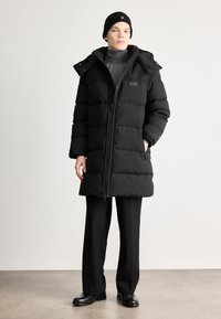 Black puffer coat with a zip front, large pockets, and a hood. Worn over a gray turtleneck and black pants, on a neutral background.